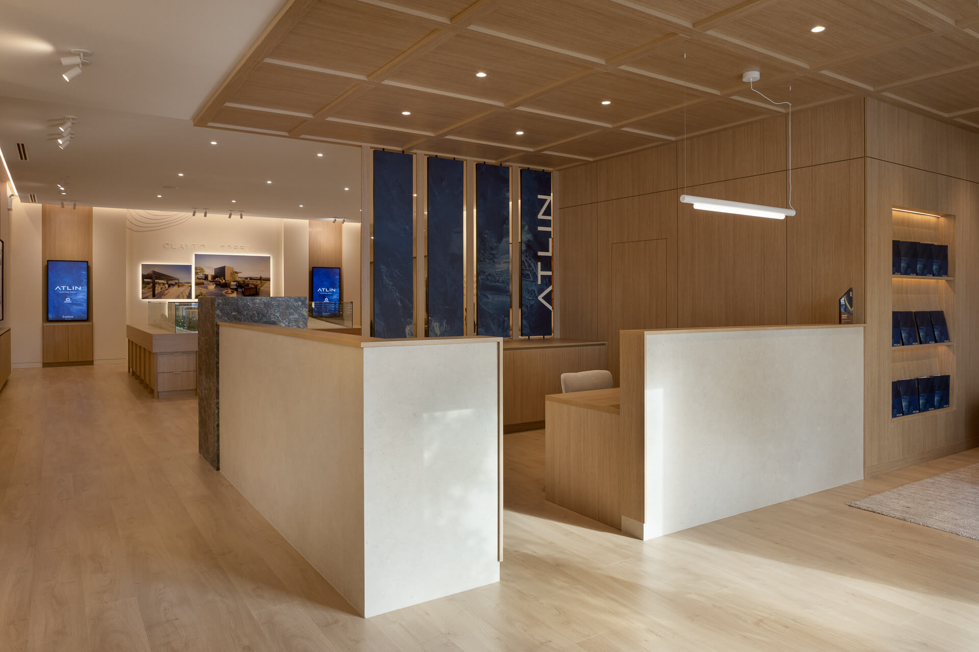 Modern luxury presentation center lobby for Atlin at Clayton Crest by Zenterra, showcasing a minimalist light-wood front reception desk, paneled ceilings, and blue branding accents