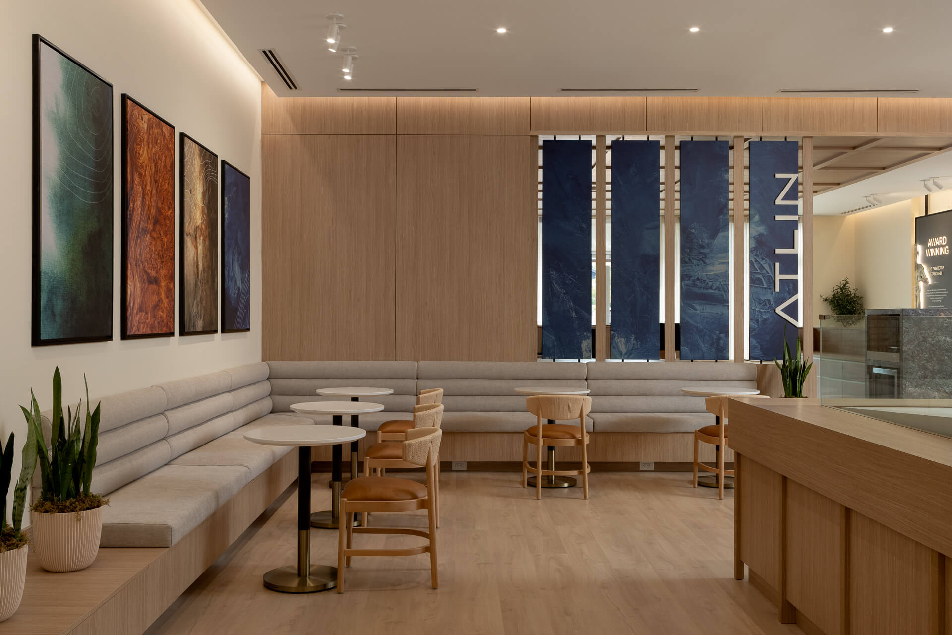 Contemporary seating lounge inside the Atlin presentation center, featuring built-in light gray banquette seating, wooden chairs, round marble tables, and vertical abstract blue wall art panels