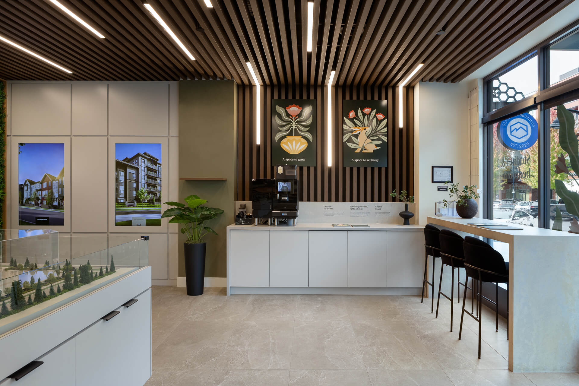 Lounge area inside Zenterra's Eden sales center, showcasing a sleek built-in white service bar with a coffee station, high-top seating overlooking the street, and decorative custom botanical posters titled A space to grow and A space to recharge