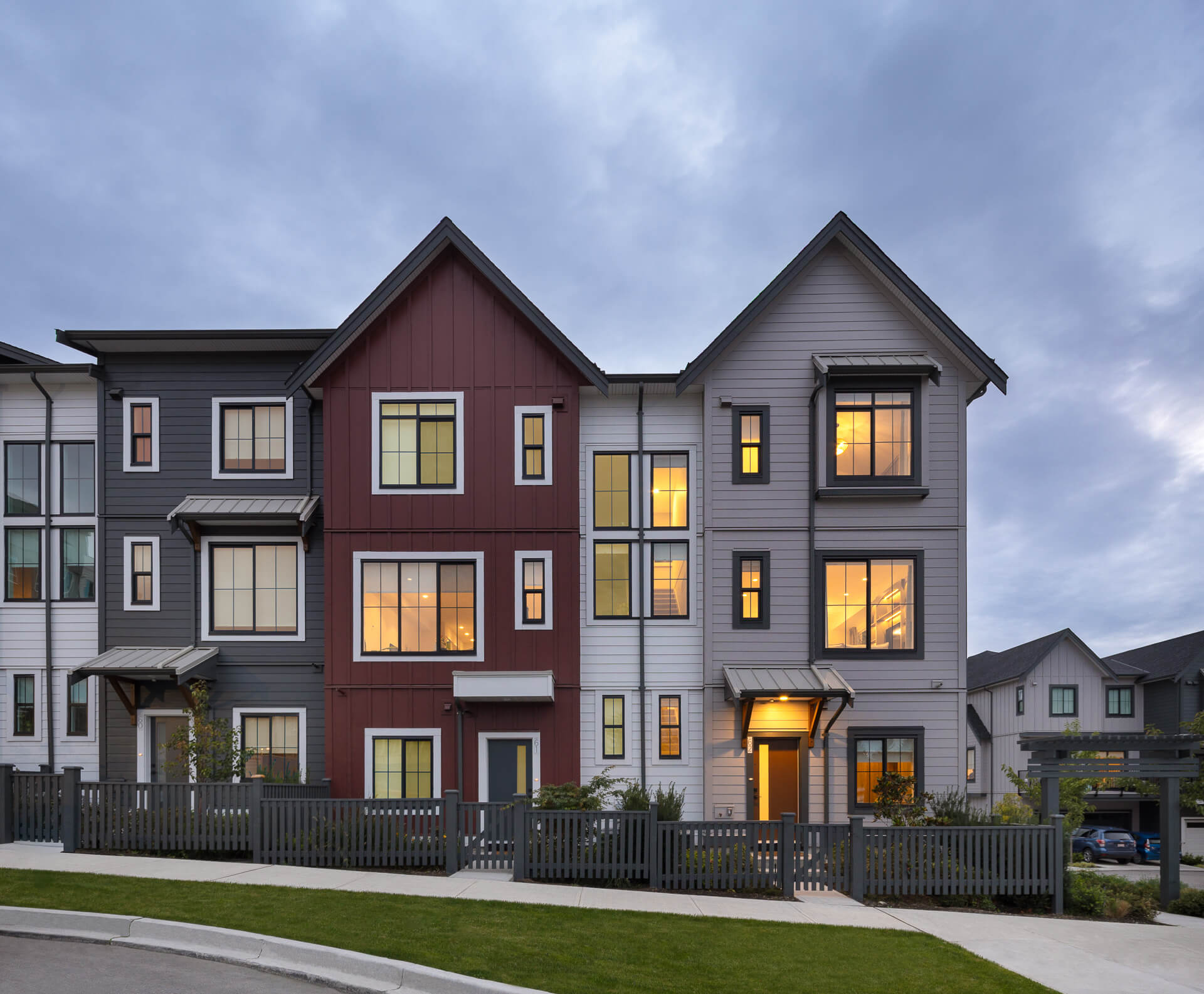 Front facade of multi-storey modern townhouses at Latimer Walk by Zenterra, showcasing contrasting dark gray, deep red, and light gray siding with illuminated windows at dusk