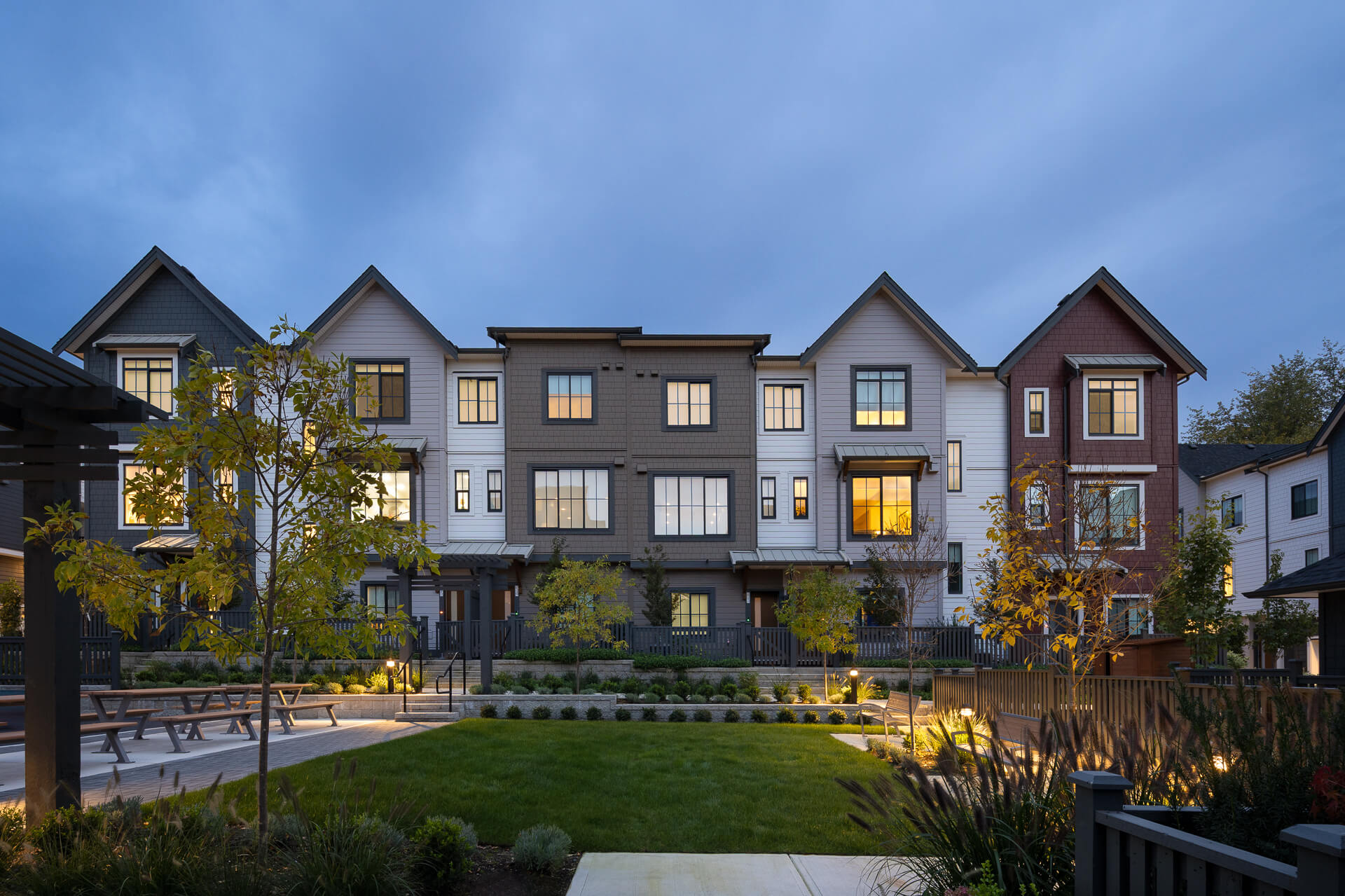 Rear view of the Latimer Walk townhome development at twilight, featuring a landscaped courtyard lawn with picnic tables and warm interior lighting glowing through the windows