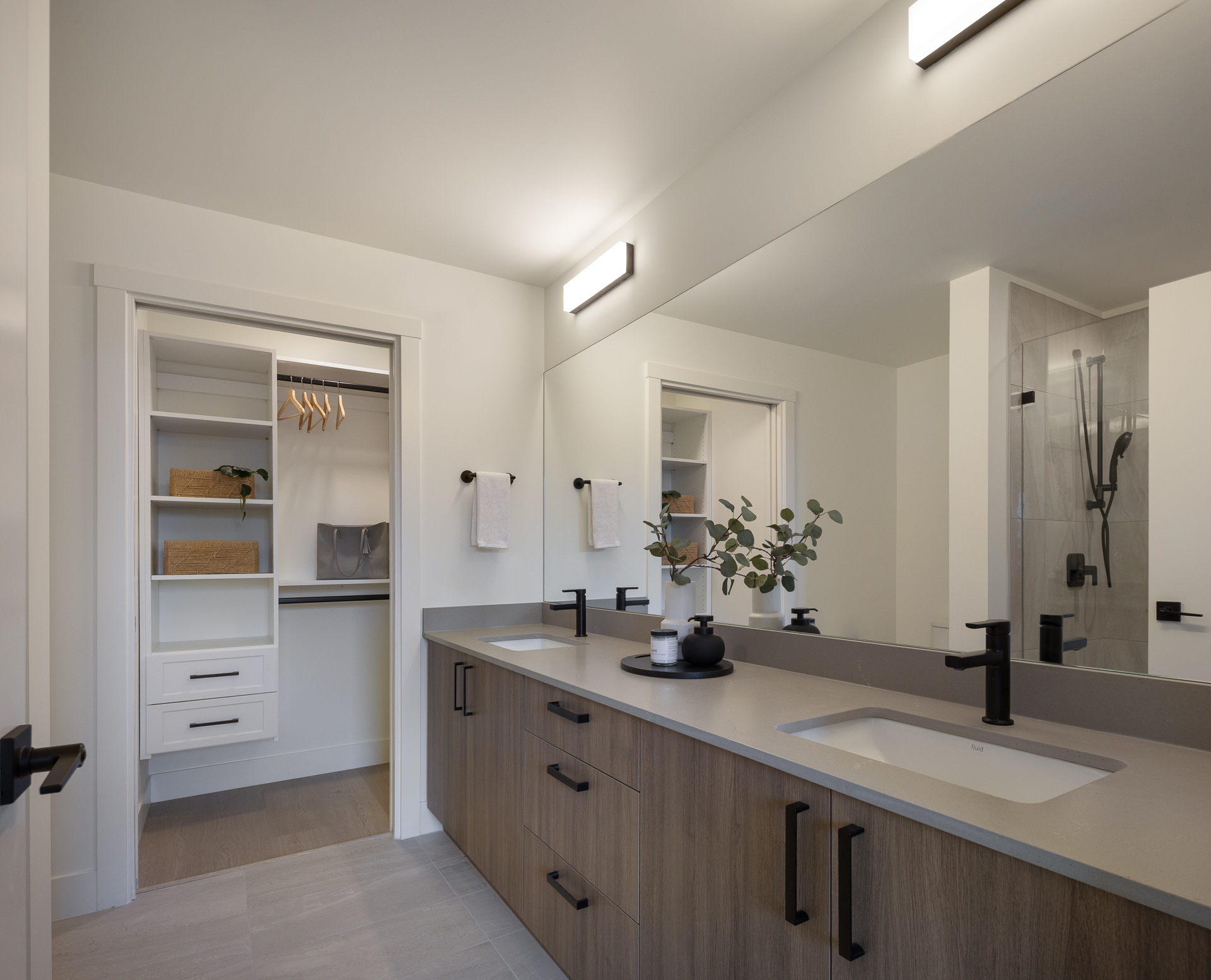 Luxury primary bathroom ensuite at The Commons condos with a double vanity, matte black faucets, large mirror, and open doorway into a walk-in closet with built-in storage organizers
