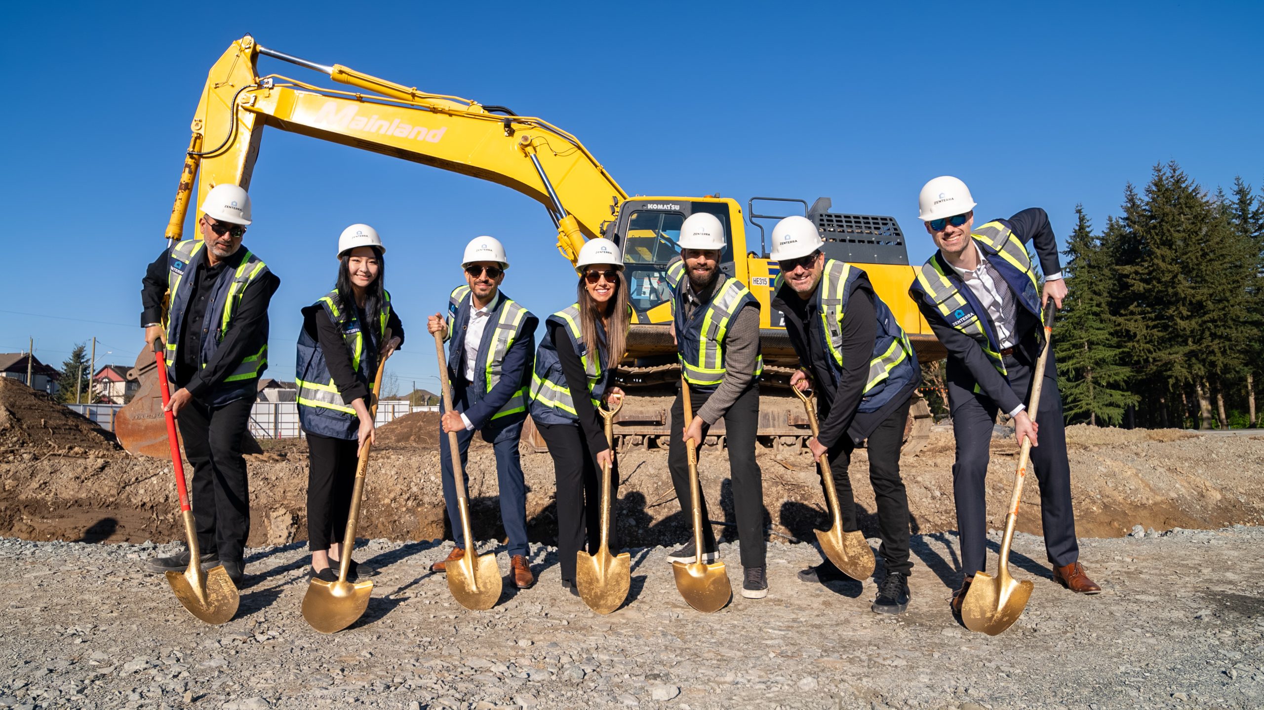 Seven members of the Zenterra Developments team wearing hard hats and safety vests while smiling and posing with golden shovels at the Clayton Crest groundbreaking ceremony in front of a yellow excavator