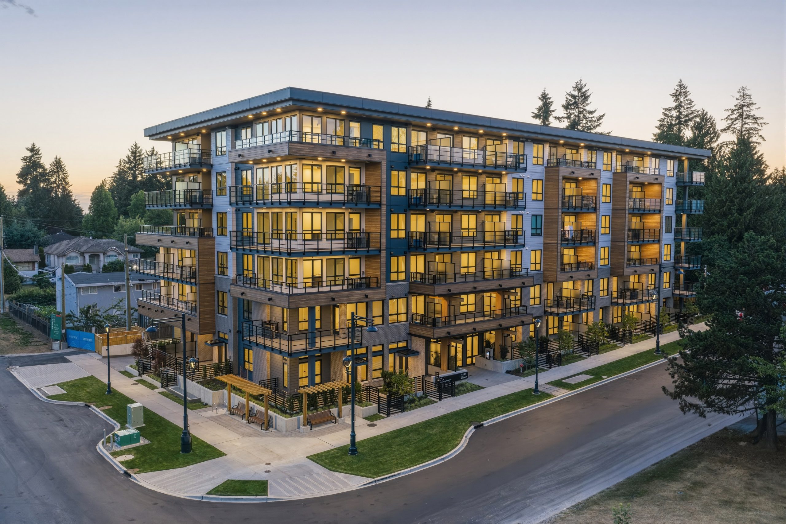 Aerial dusk view of the City Corners condominium building by Zenterra Developments, showcasing modern wood-toned cladding, grey paneling, large glowing glass windows, and landscaped corner pathways