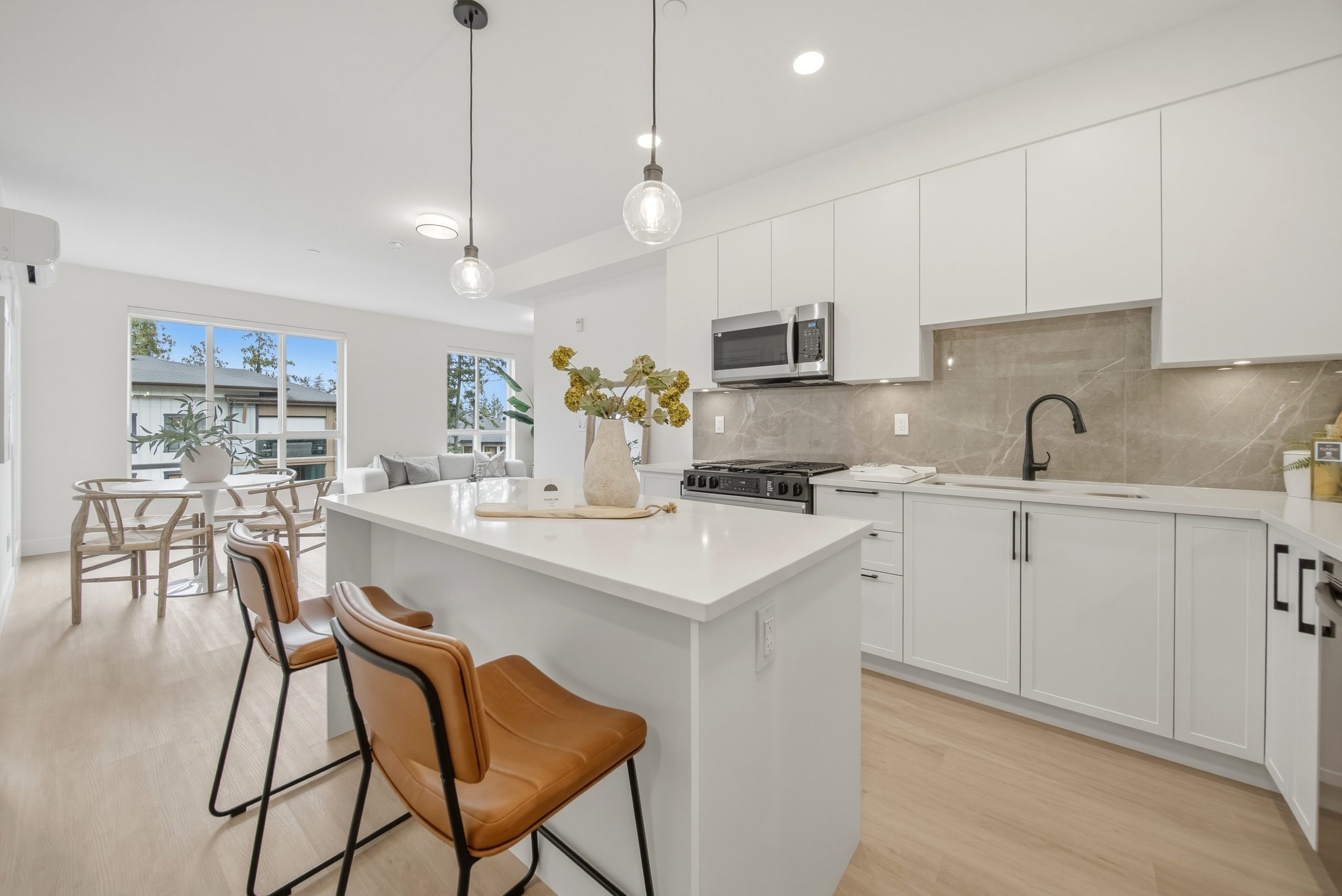Detailed photograph of a gourmet kitchen within a King and Crescent residence. The space showcases a large white quartz island with cognac-brown leather bar stools. Standard features, exemplifying the quality of this South Surrey project, include full-height glossy white flat-panel cabinets, a light grey slab backsplash, and high-performance stainless steel appliances. Large windows provide natural light and views of a treed, park-like neighborhood.