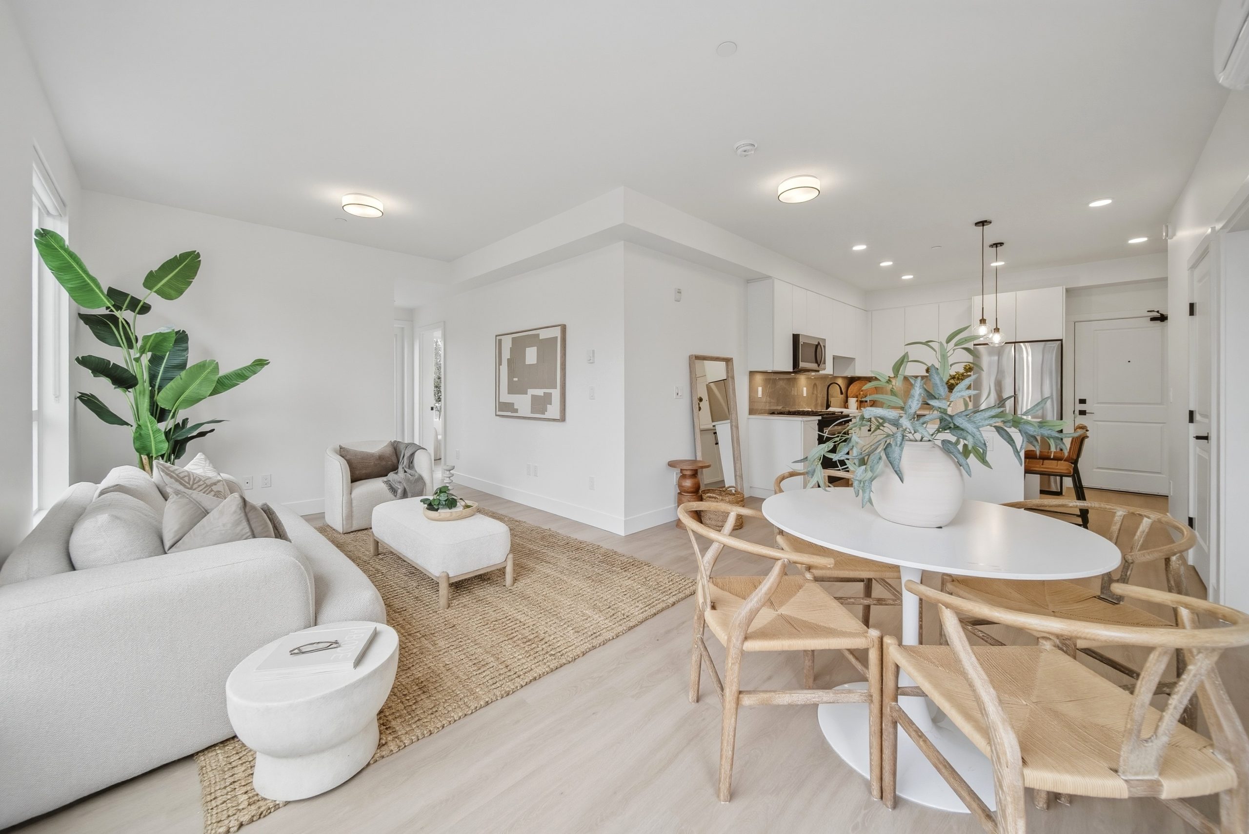 Interior view of a contemporary, open-concept display suite at King and Crescent by Zenterra. The design features wide-plank wood flooring and pristine white walls. In the foreground is a dining table with wishbone-style chairs. The living area, viewed here as an example of Zenterra’s higher standard of finishing, includes a large cream-colored sectional sofa and a natural jute rug, leading toward private balconies.