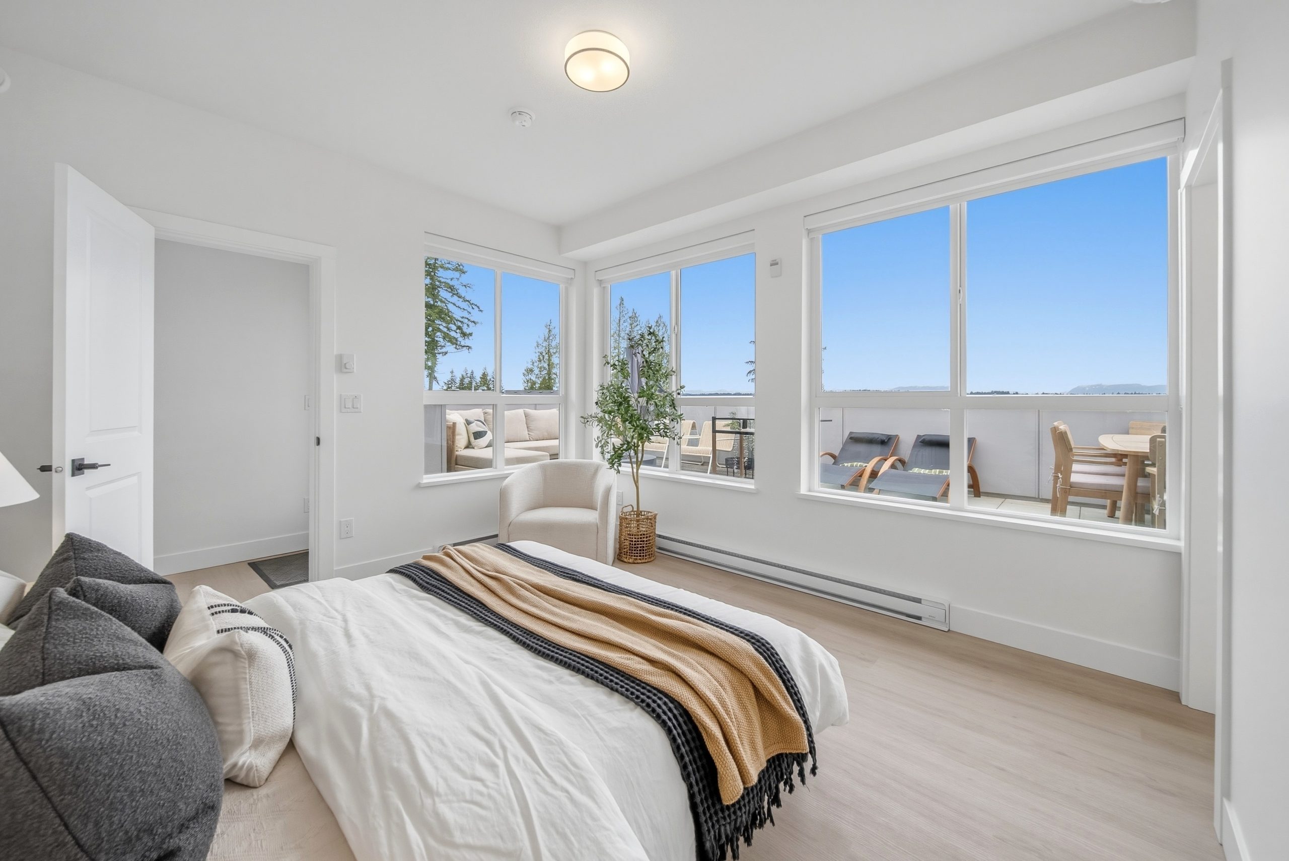 A spacious primary bedroom interior within the King and Crescent project by Zenterra, featuring light-oak wide-plank flooring and minimalist styling. The bed is layered with textured linens and substantial grey pillows. Large, black-framed corner windows stretch across two walls, flooding the space with daylight and offering direct access to integrated private balconies that overlook the surrounding green spaces and trees.