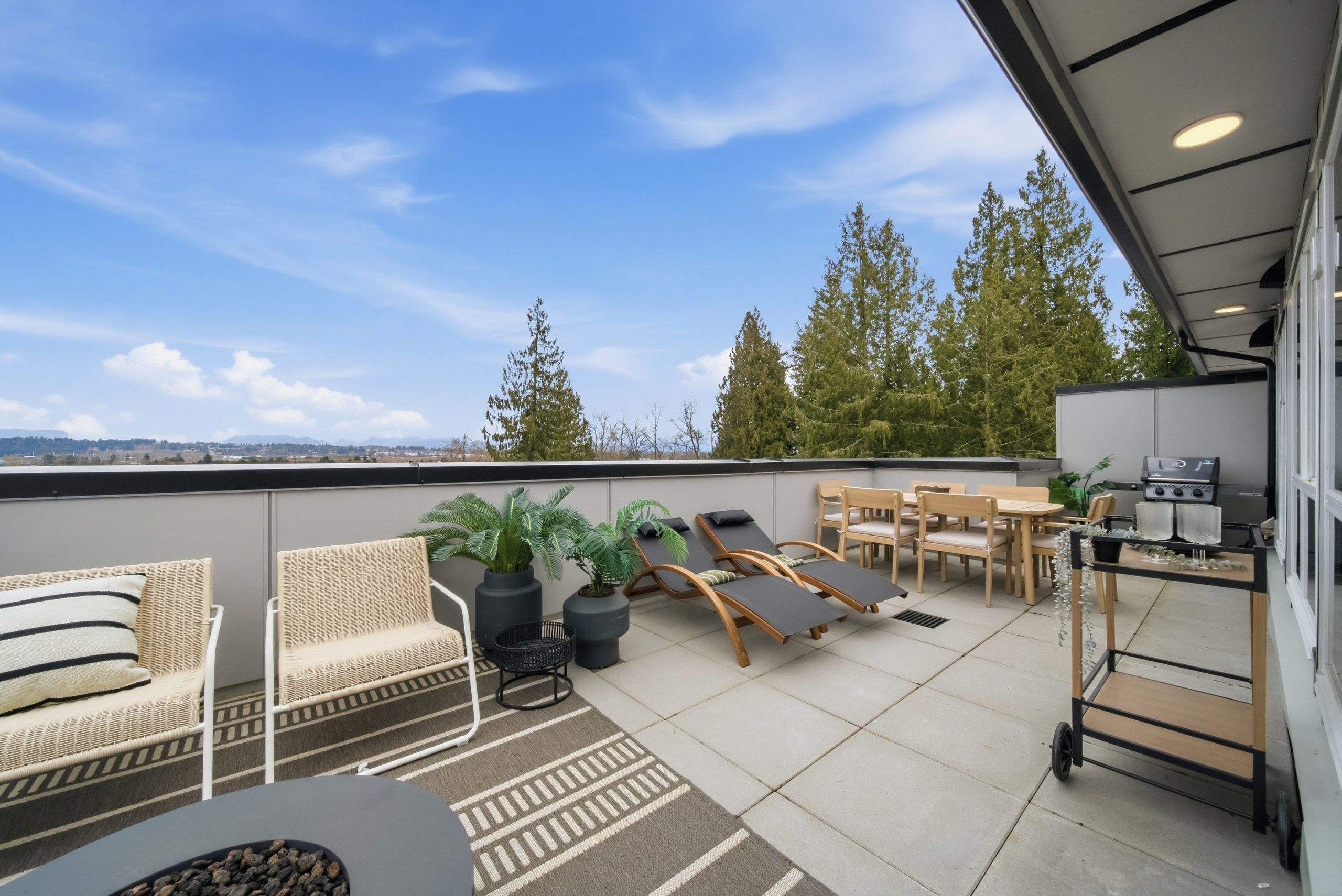 Extensive view of the integrated private balconies attached to a suite at King and Crescent by Zenterra. This large outdoor space, corrected here as expansive balconies rather than a rooftop, is paved with concrete pavers and features distinct functional zones: a cozy conversational area with woven chairs around a round gas fire table, a sun-lounging area with teak chaise lounges, and a full teak dining set with seating for six. The balconies are framed by tall evergreen trees under a bright blue sky.
