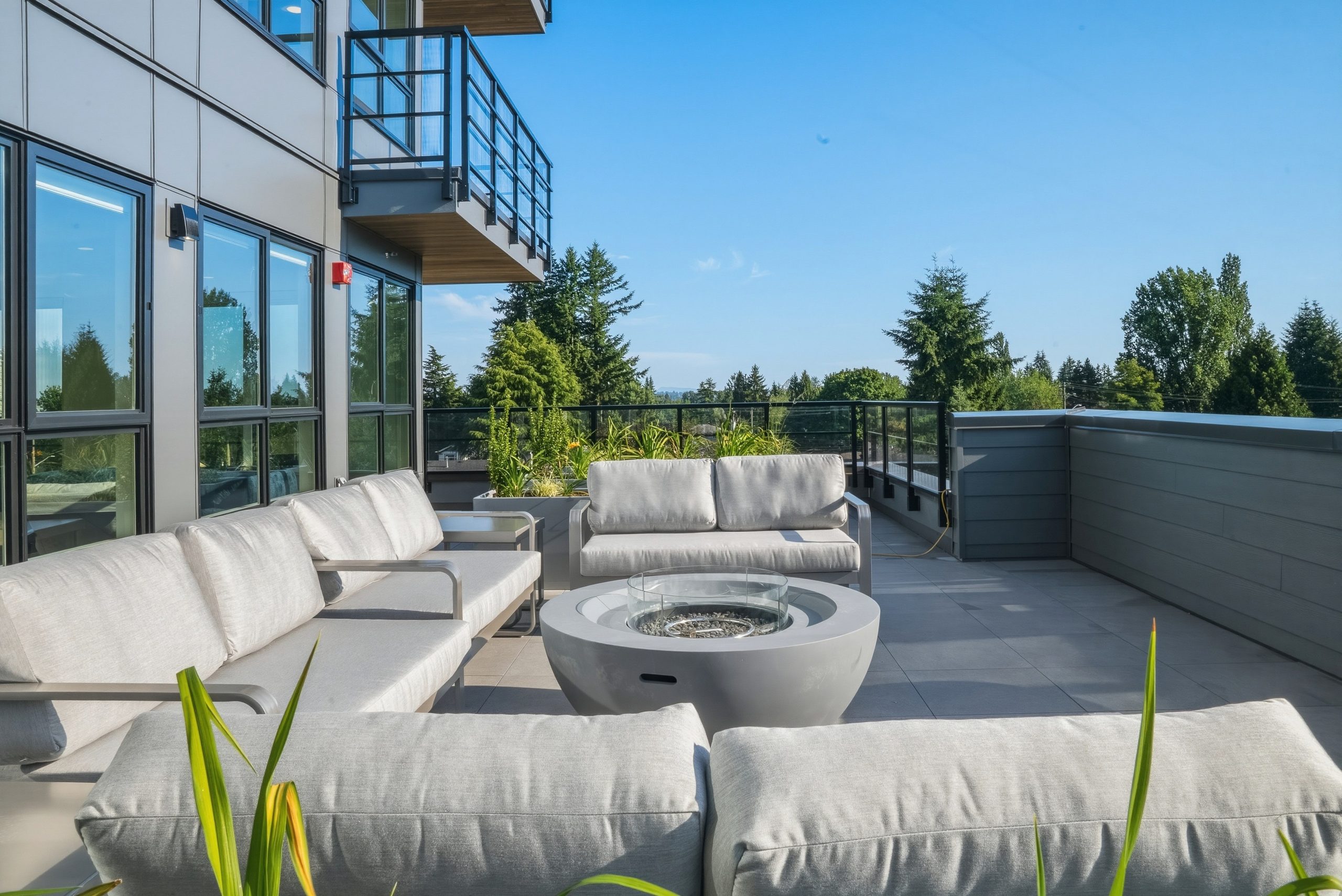A sunny rooftop terrace lounge at City Corners by Zenterra, equipped with comfortable grey sectional outdoor sofas, a central concrete fire bowl, and views overlooking treetops