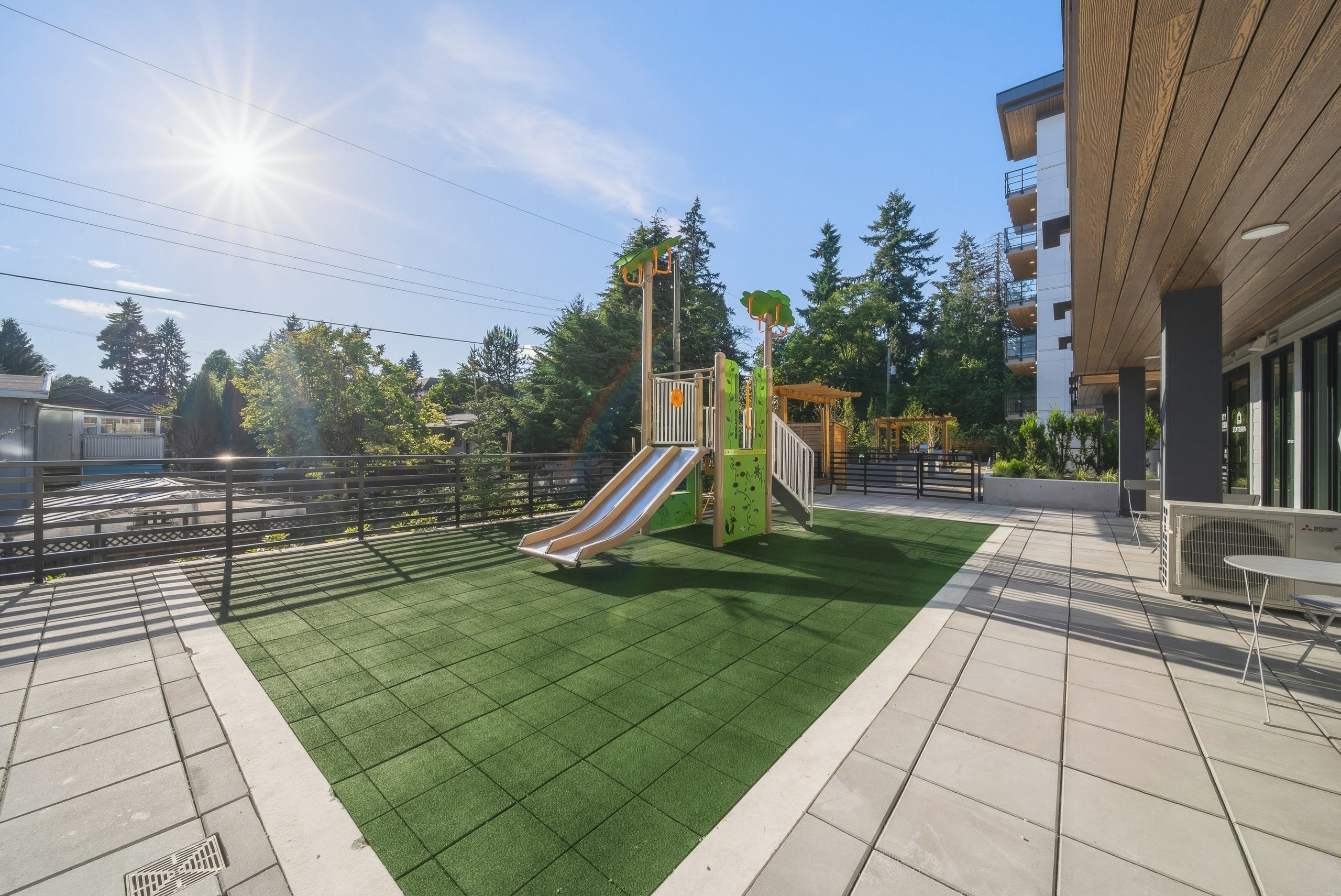 The community children's playground area at City Corners, featuring a green artificial turf lawn, a small play structure with slides, a wooden arbor, and bench seating