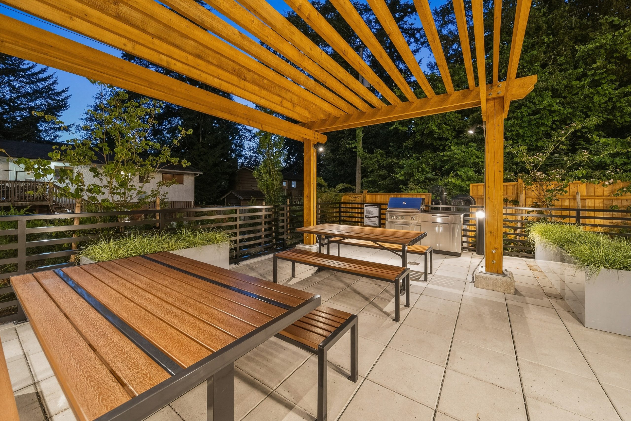 An outdoor common amenity area at City Corners featuring a heavy timber wooden pergola over long picnic tables and a built-in stainless steel outdoor kitchen BBQ setup at twilight